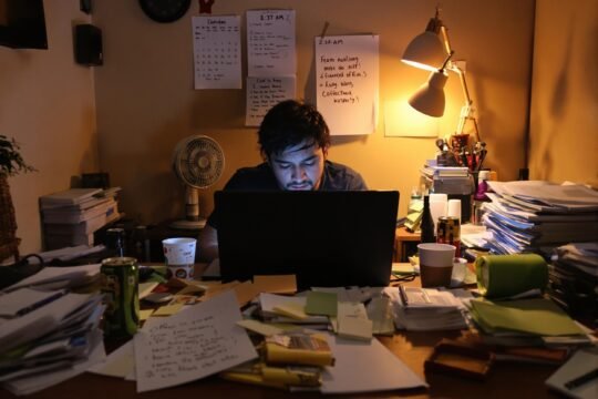 Pria muda Indonesia duduk lelah di meja kerja kamar gelap, wajah bingung dan kosong, menggambarkan hidup yang sibuk tapi terasa tidak bergerak ke mana-mana.